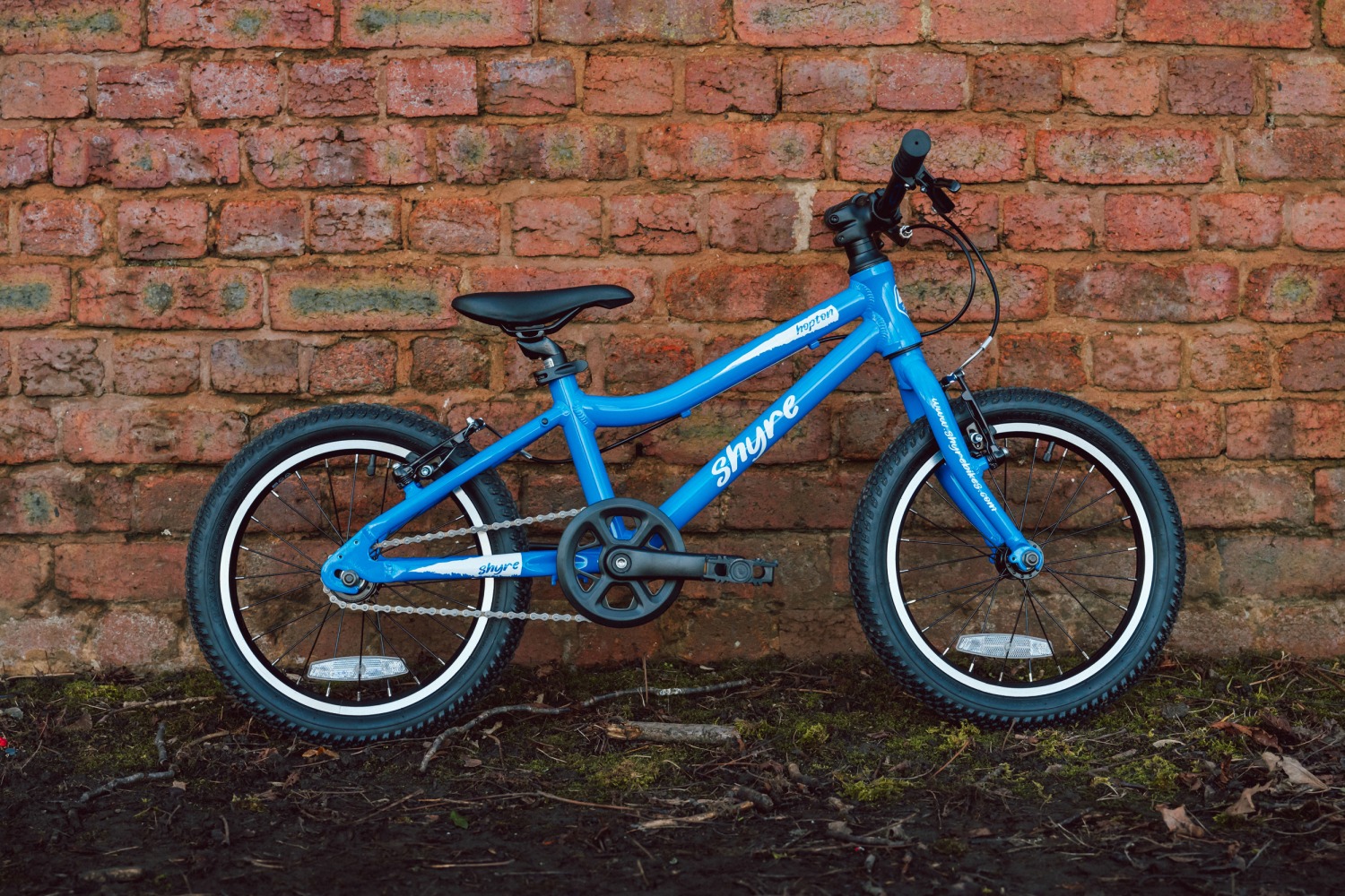 Adam LV Photography_ Shyre bikes Shoot static-71 A blue Shyre bikes Hopton 16 bike seen from the side, leaning against a brick wall