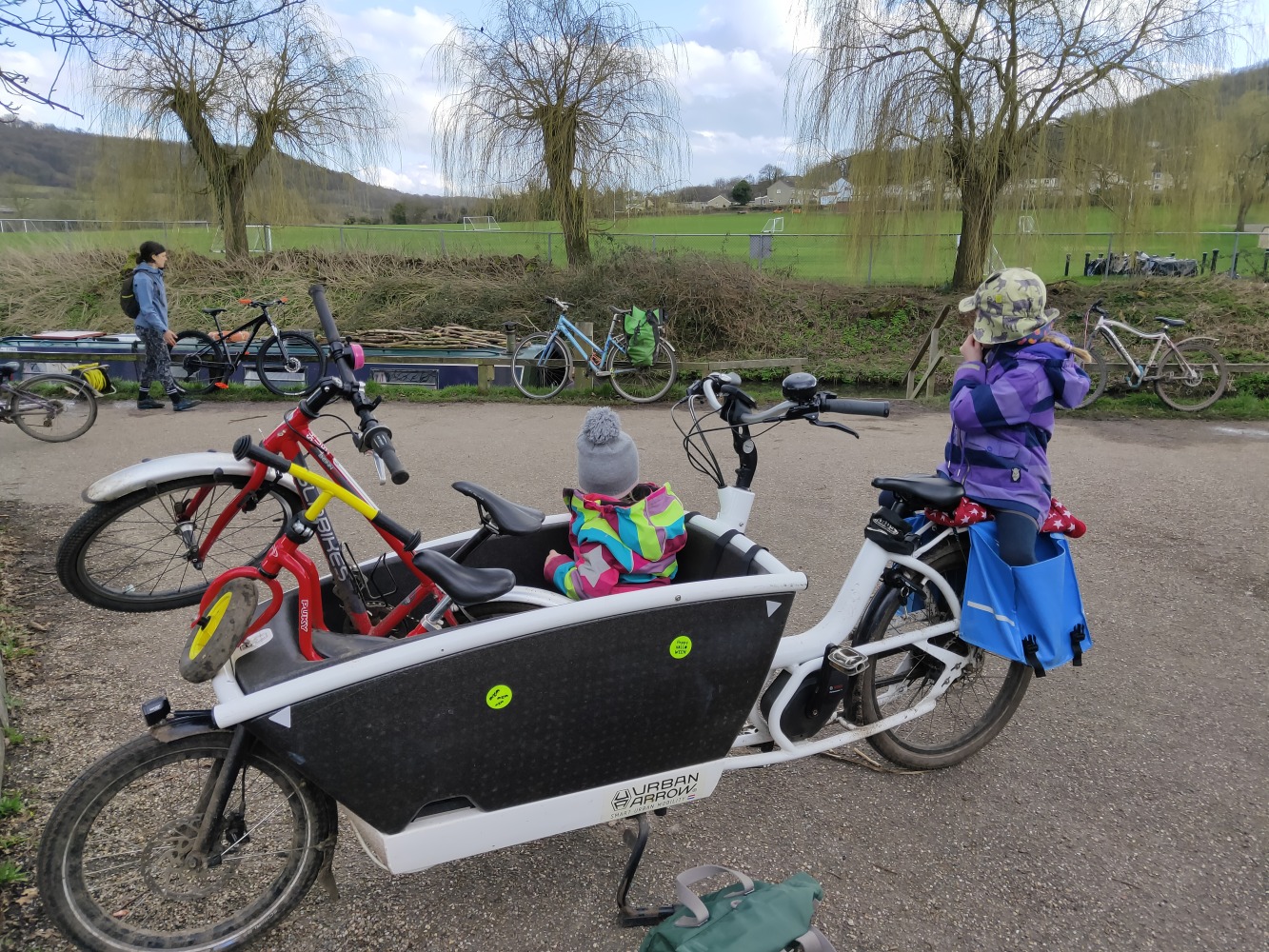 Urban Arrow Family loaded with a kids bike and a balance bike and a child sitting on bench