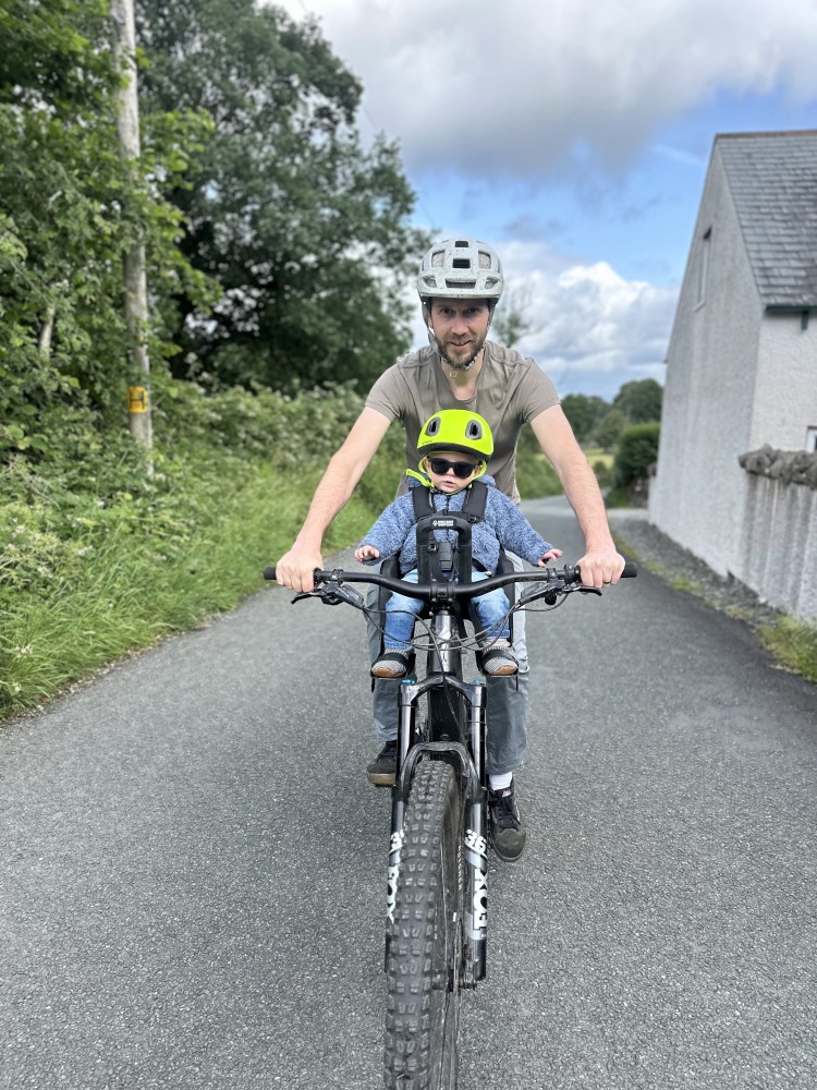 A little boy with a yellow helmet and sunglasses smiling on the kids ride shotgun pro evo seat with his dad riding on a tarmac road