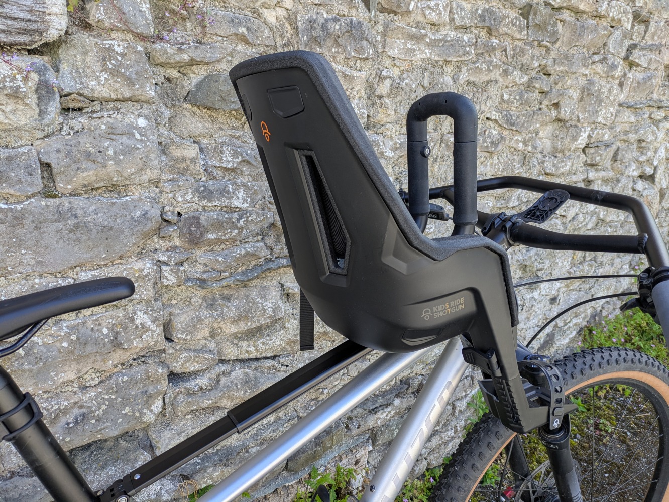 A Kids Ride Shotgun Pro Evo reat mounted to a silver mountain bike leant against a brick wall. a close up of the black plastic seat