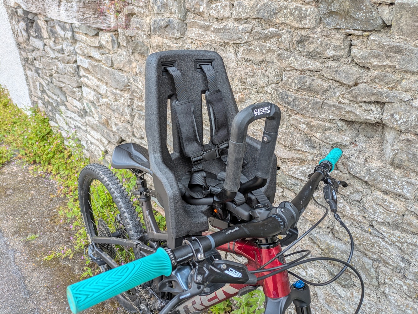 A Kids Ride Shotgun Pro Evo baby bike seat mounted to a red mountain bike leant against a stone wall
