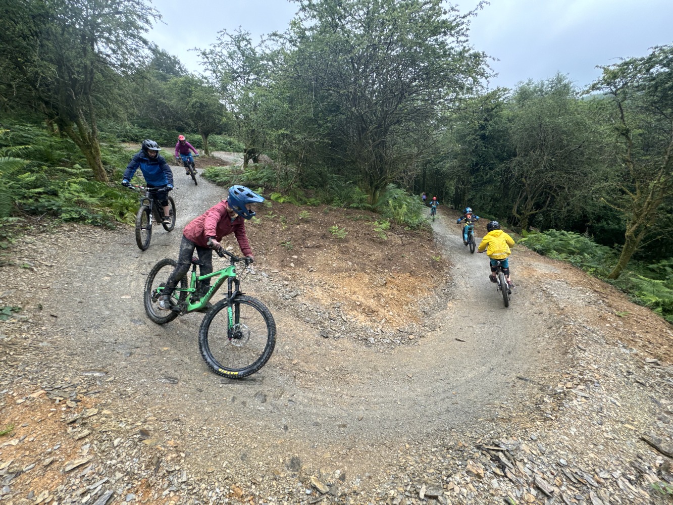 Charlie Kingston – hafod A group of mountain bikers, including kids, riding round a gravelly berm, surrounded by trees