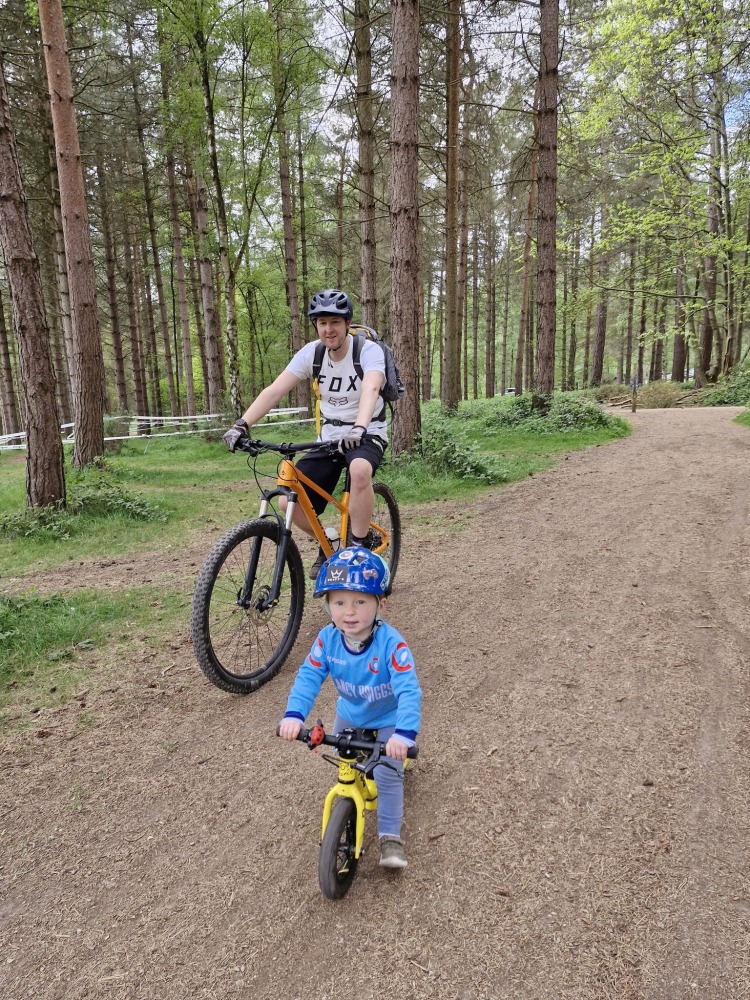 Charlotte Quinn 1 – sherwood A man riding a mountain bike alongside a small boy on a balance bike, with woodland in the background, seen from the front