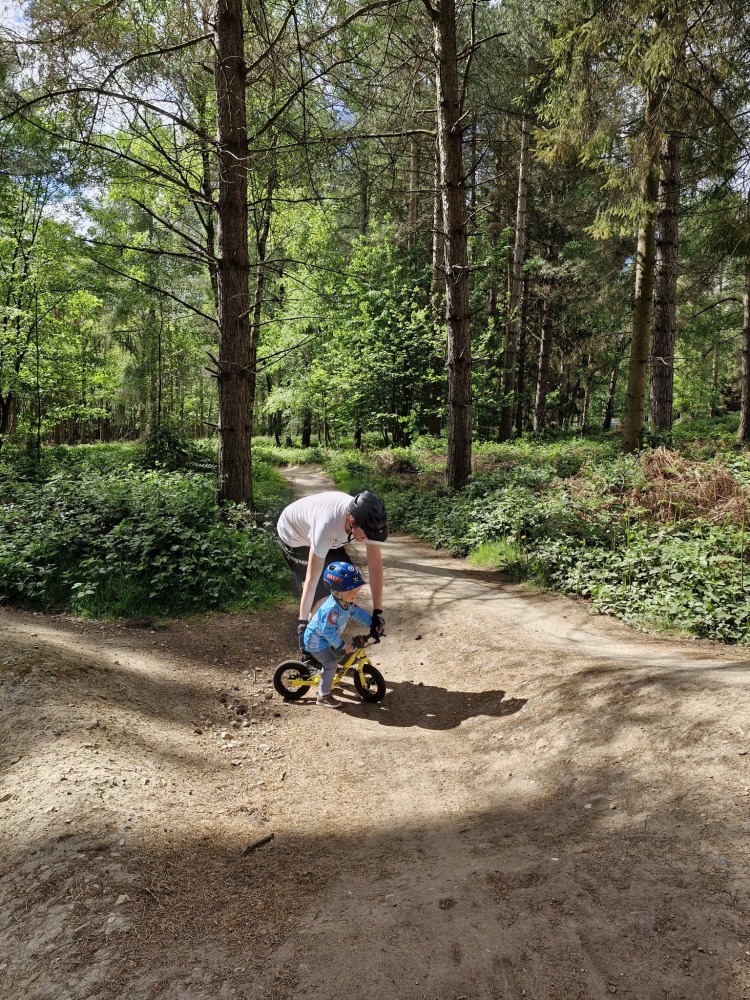 Charlotte Quinn 2 – sherwood A dad bending over and helping his small son on his balance bike, in the middle of the woods