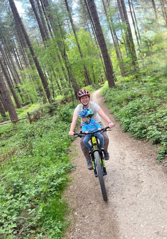 Charlotte Quinn 3 – sherwood A woman seen from the front, riding a mountain bike through woodland, smiling at the camera, with her young son on a front bike seat