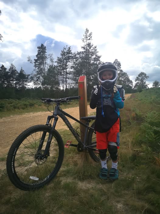 Conor Ford – Swinley A little girl wearing a full face helmet standing next to her mountain bike and a red trail sign post, giving two thumbs up to the camera