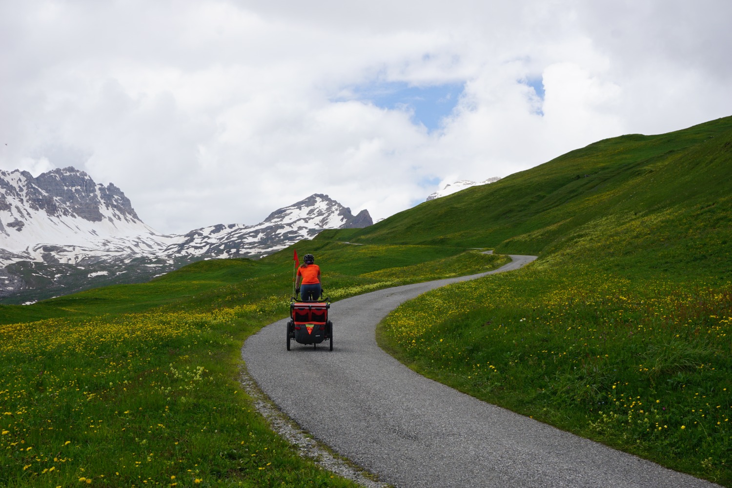 Copy of DSC02145 Hetty Kingston seen from behind cycling with the trailer along a smooth winding tarmac path with Swiss snow-topped mountains in the distance