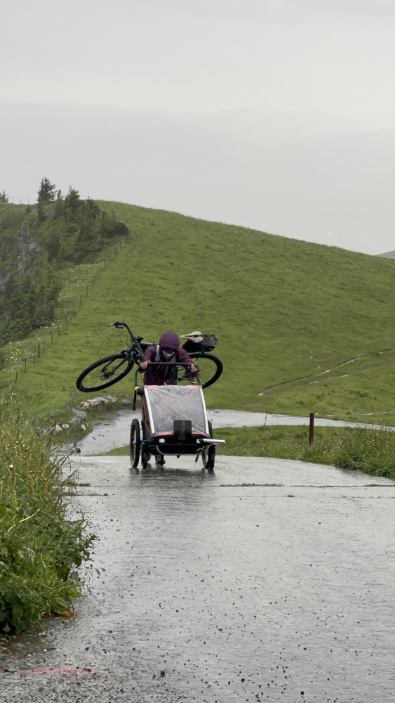 Copy of IMG_6757 Hetty Kingston seen from a distance, carrying a bike on her back and pushing a kids trailer along a wet, smooth road
