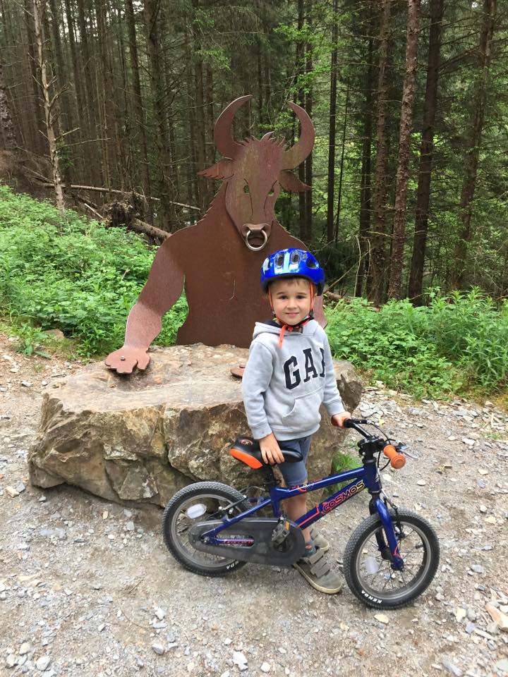 Debbie Kojs – CYB 4 A little boy wearing a blue helmet posting with his bike in front of a Minotaur sign in the woods