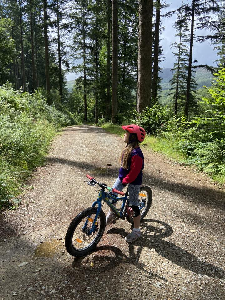 Debbie Kojs – CYB A little girl standing over her mountain bike on a gravel path surrounded by woodland, looking up at the trees