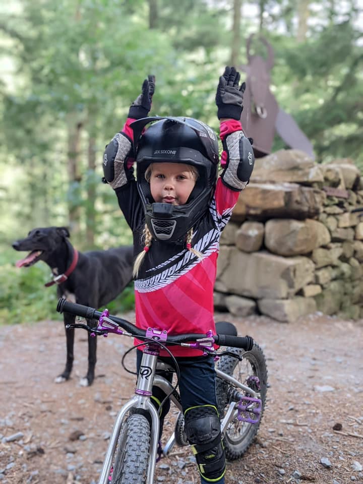 Laura Rees – CYB A little girl wearing a full face helmet, standing over a little bike and holding her hands in the air, with a dog and a Minotaur sign in the background