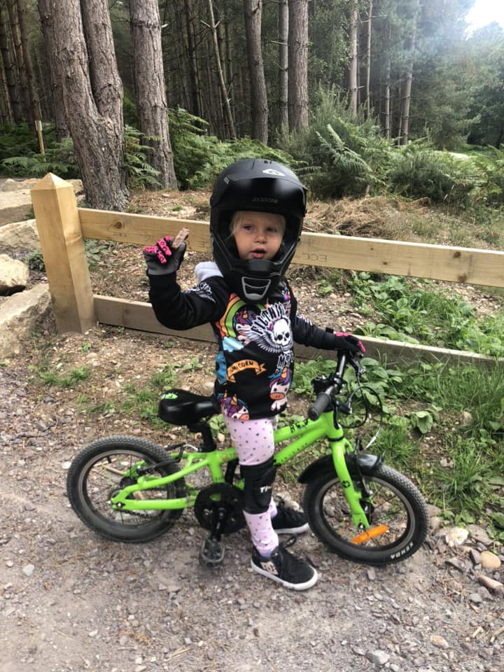 Laura Rees – Cannock 2 A little girl in a full face helmet on her green bike, holding a snack up to the camera