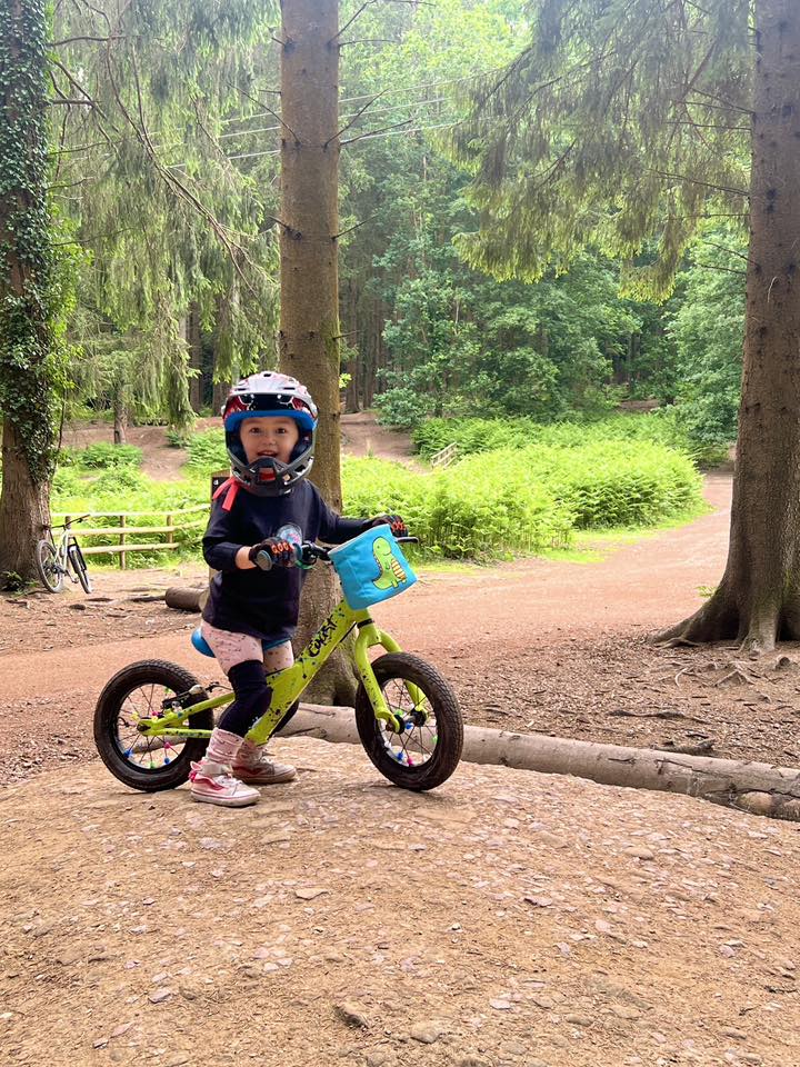 Rebecca Prayoga 2 FoD A little girl on a yellow balance bike and wearing a full face helmet, smiling among some trees
