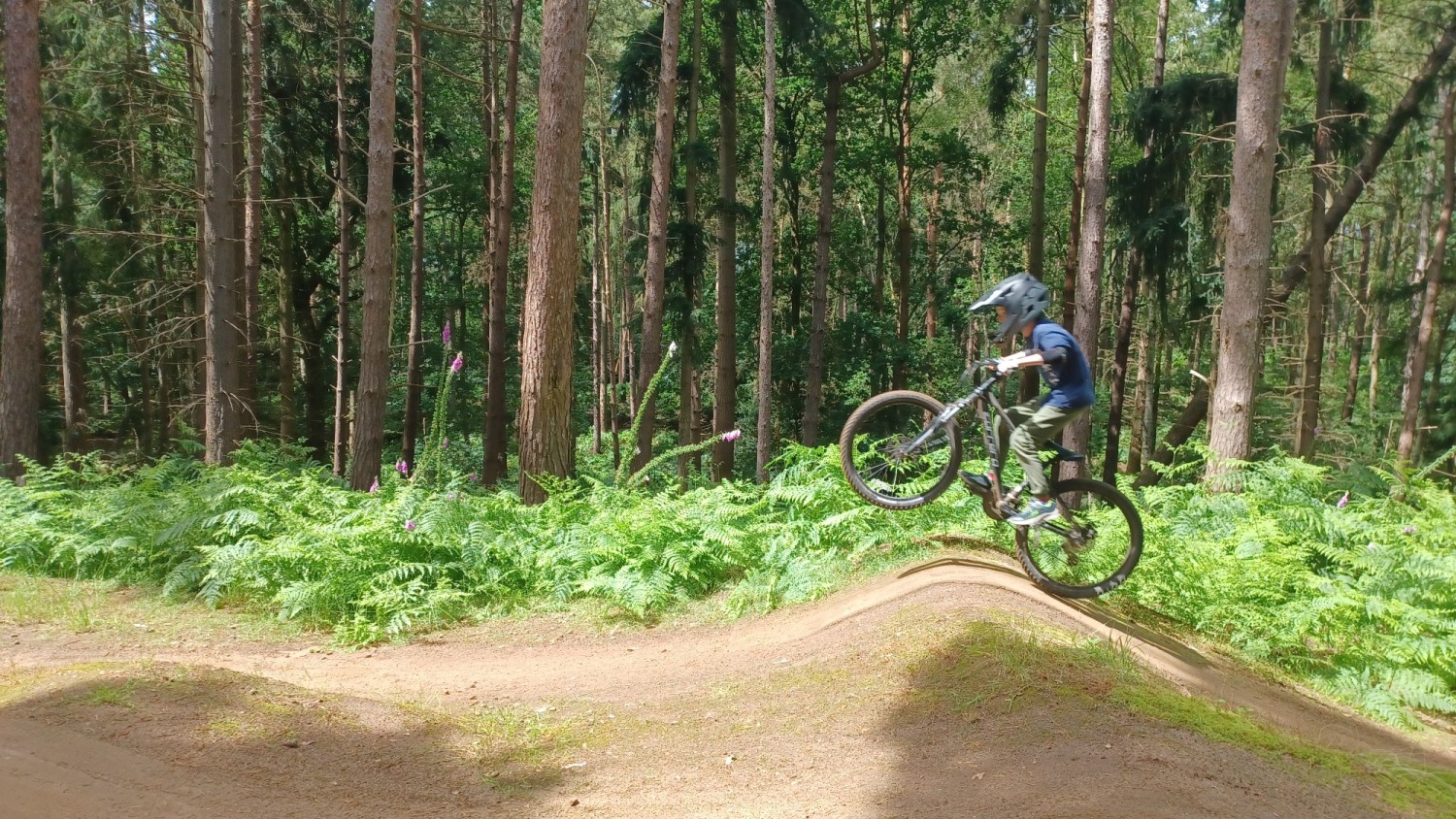 Ruth Preece – Chicksands A small child wearing a full face helmet and manualling their small mountain bike over the crest of a jump with woodland behind them