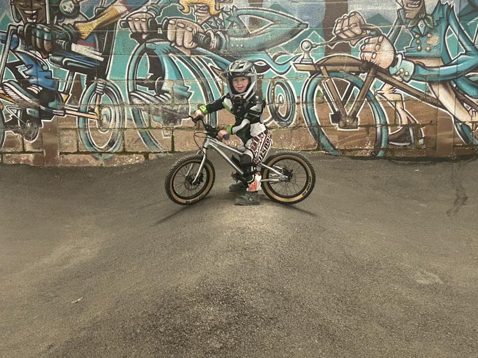Sarah McWilliam-Free – Bike Park 417 A very young girl standing on a pump track in front of a graffiti wall, over a small pedal bike, wearing a full face helmet and smiling