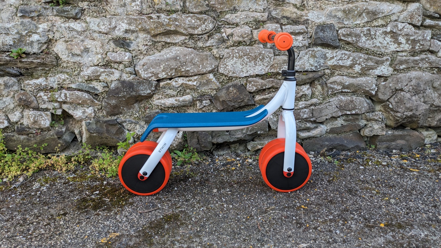 Btwin Convertible 2-in-1 Ride-On to Balance Bike, a white, orange and blue bike against a stone wall