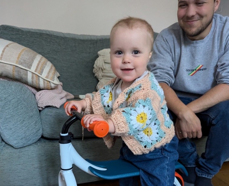 A little girl in a flowery cardigan sat on the Decathlon 2-in-1 ride-on baby balance, a white, orange and blue bike in her living room