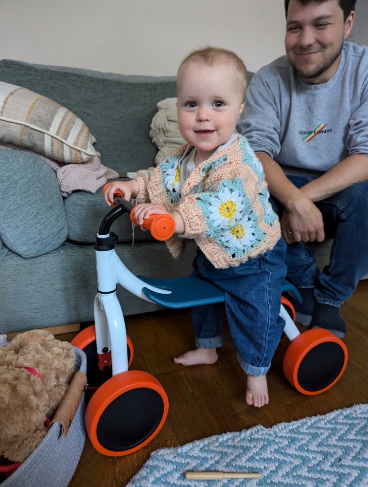A little girl in a flowery cardigan sat on the Btwin Convertible 2-in-1 Ride-On to Balance Bike, a white, orange and blue bike in her living room