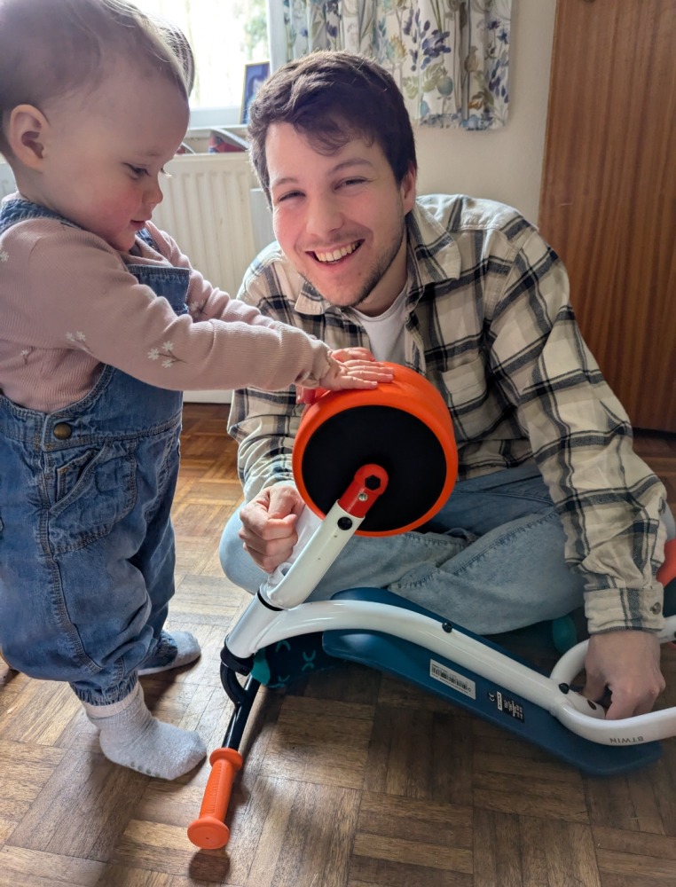 Decathlon 2-in-1 ride-on baby balance bike A little girl in dungarees is looking at the wheels on the Decathlon 2-in-1 ride-on baby balance, a white, orange and blue bike with her dad