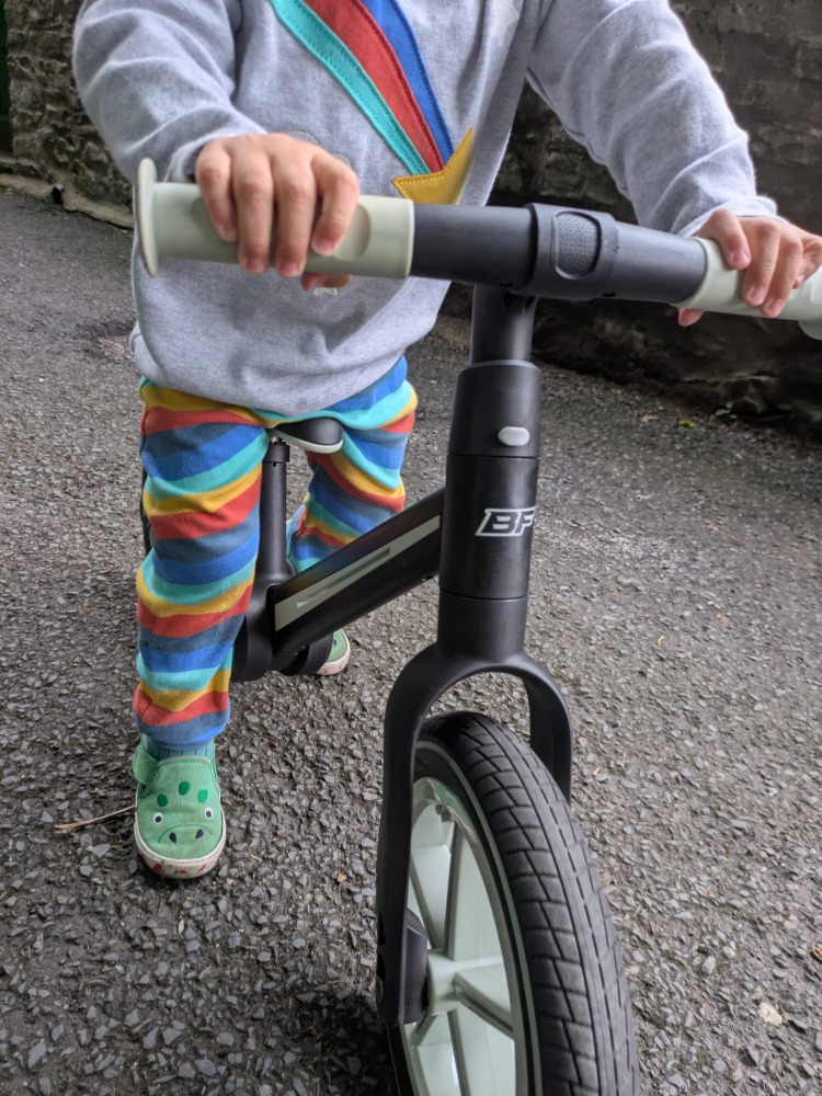A little boy wearing stripy leggins and a coulourful helmet riding the blue and black Be Fun wheels folding balance bike, a close up of the grips