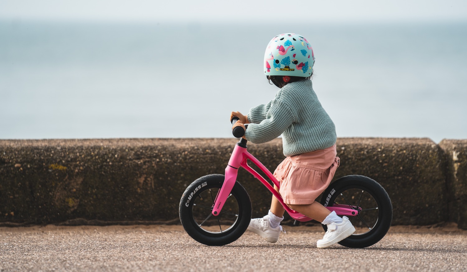 A toddler wearing a pink dress and blue cardigan riding a pink Hornit AIRO 12 balance bike along the sea wall on the coast