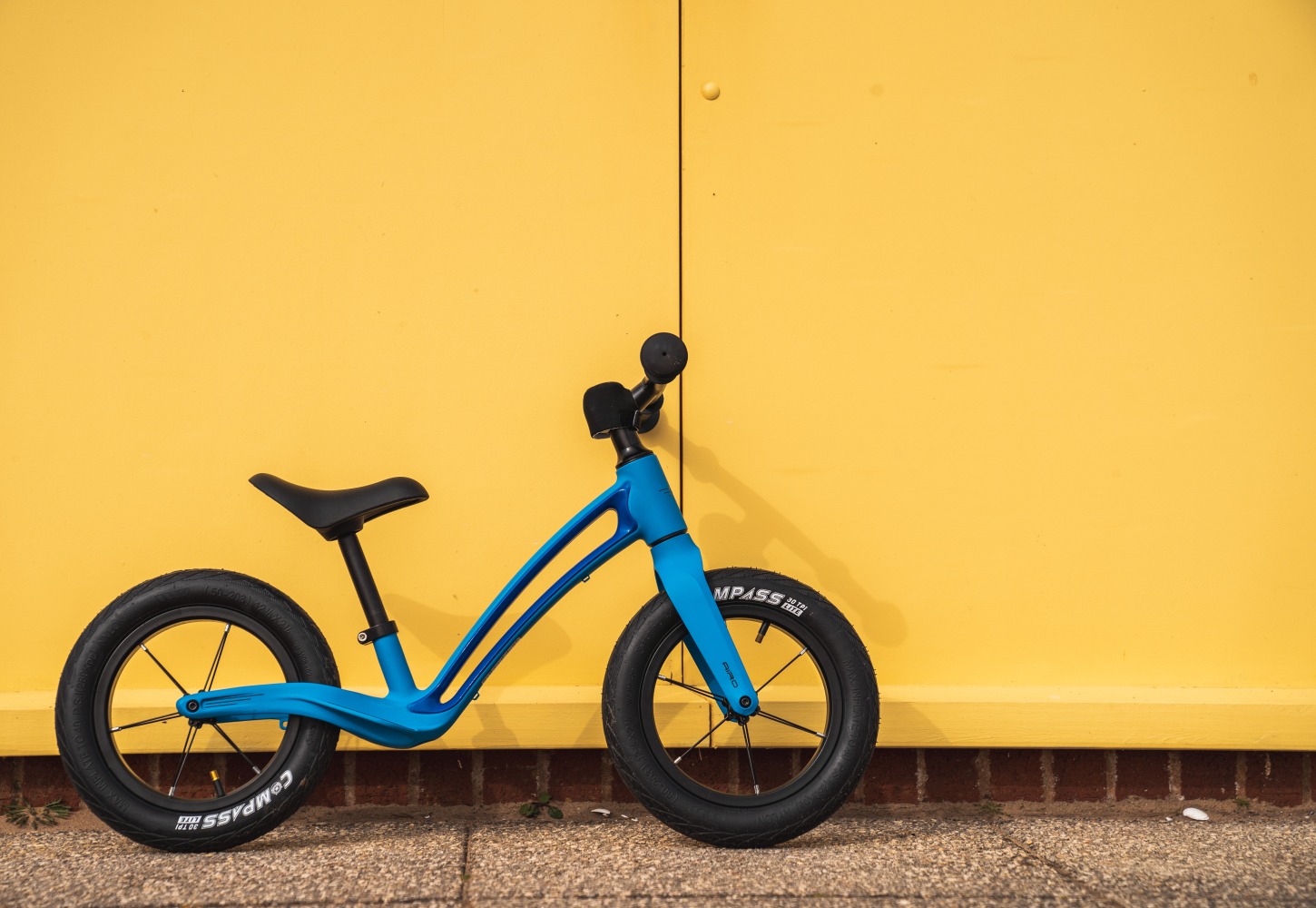 A blue Hornit AIRO 12 balance bike leaning against a yellow wall