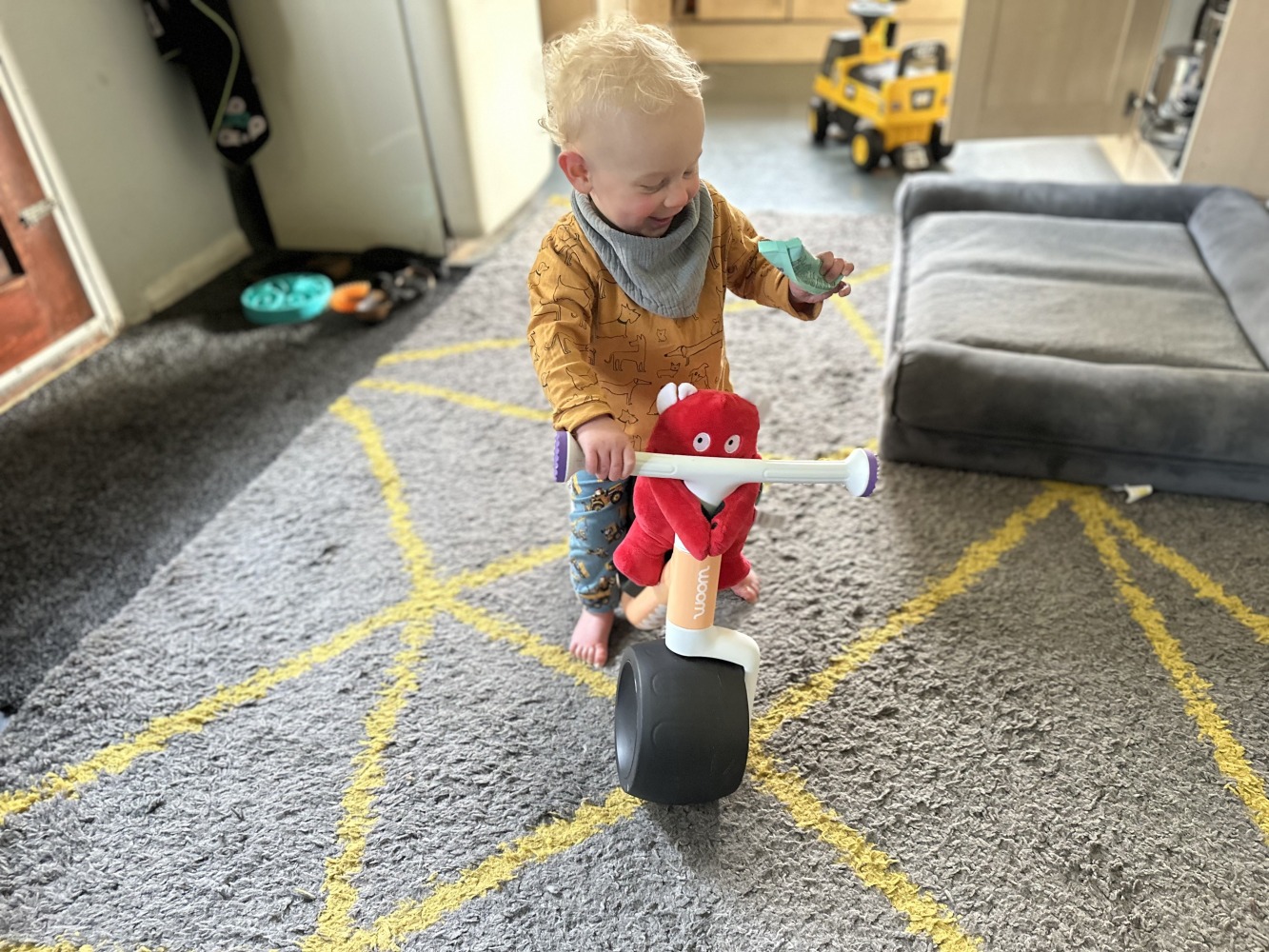 Toddler riding the woom WOW balance bike inside with Buddy the soft toy mascot on the handlebars