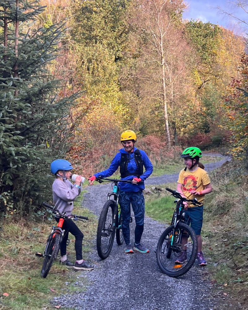 A girl wearing a blue SPARKY helmet- riding her orange mountain bike with her mum and sister