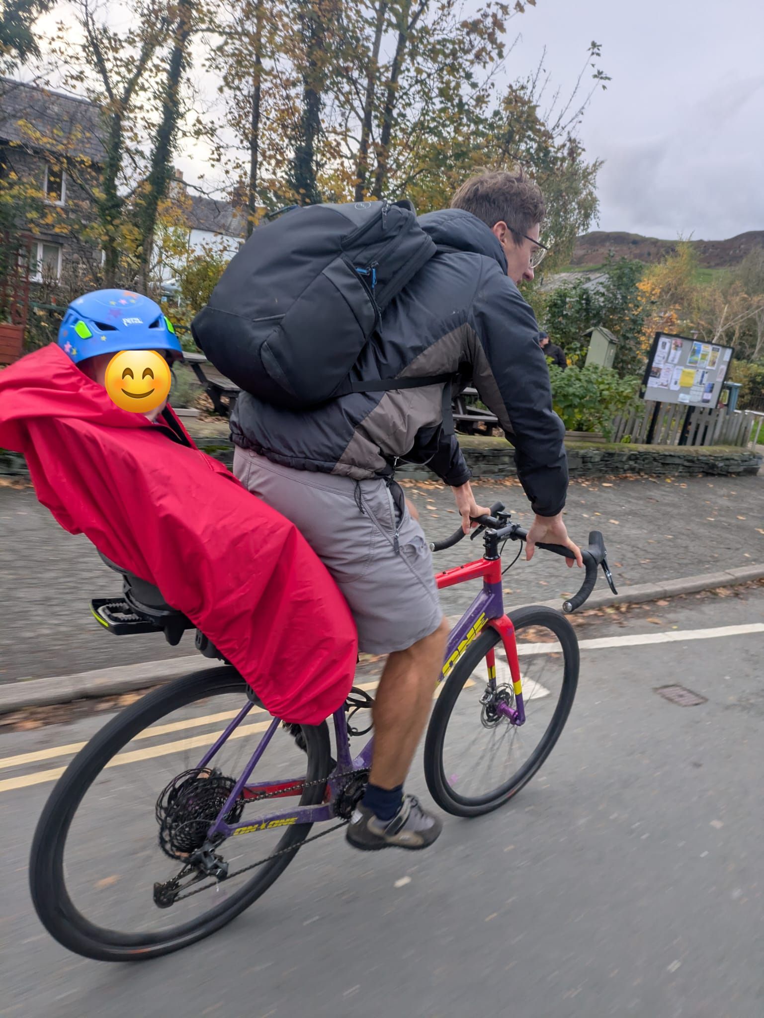 a little boy on a rear bike seat in his red Hamax Bike Seat Rain Poncho on his dads bike