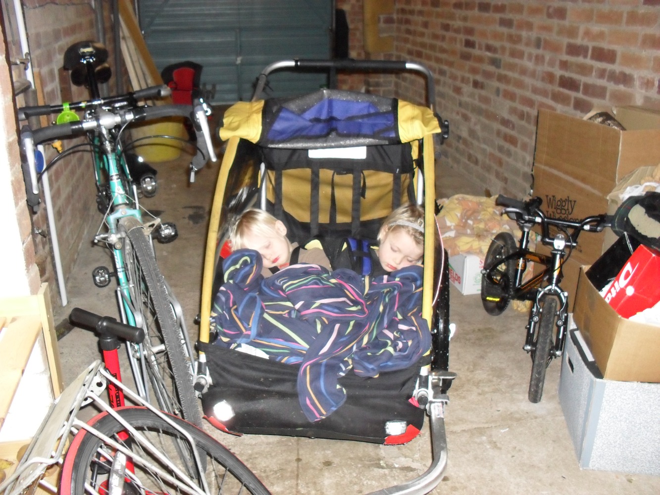Two small blonde children fast asleep in a bike trailer, with a stripy fabric baby carrier for a blanket. The trailer is in a brick garage with a closed door, attached to a bike that’s leaning sideways and next to a turquoise tandem. A small 14in wheel pedal bike is leaning on cardboard boxes nearby.