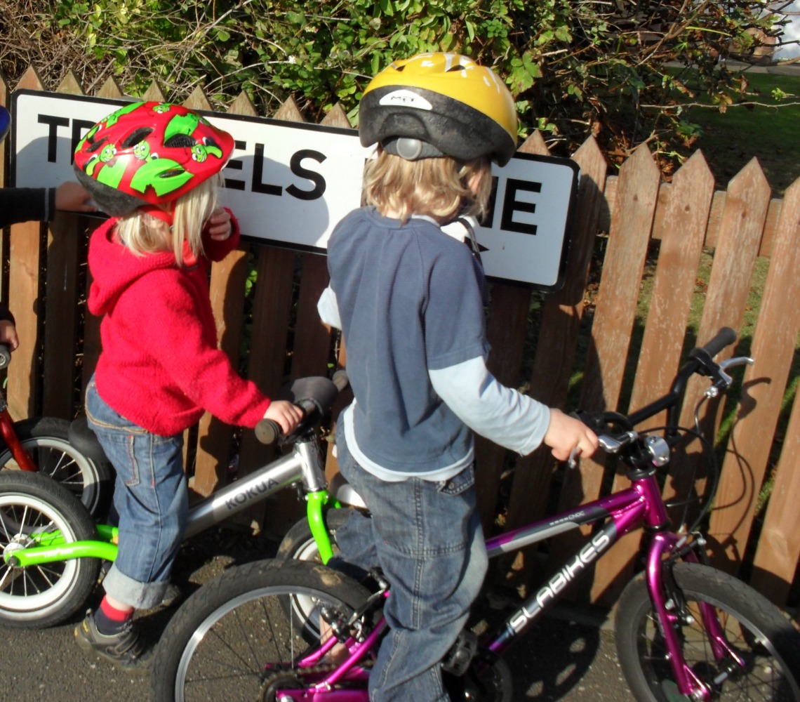 Kid1 on a pedal bike and Kid2 on a balance bike intensely reading a street sign. Another child’s is just in view to the left, and the front wheel of another balance bike – I was a childminder and often out with four small children and four small bikes!