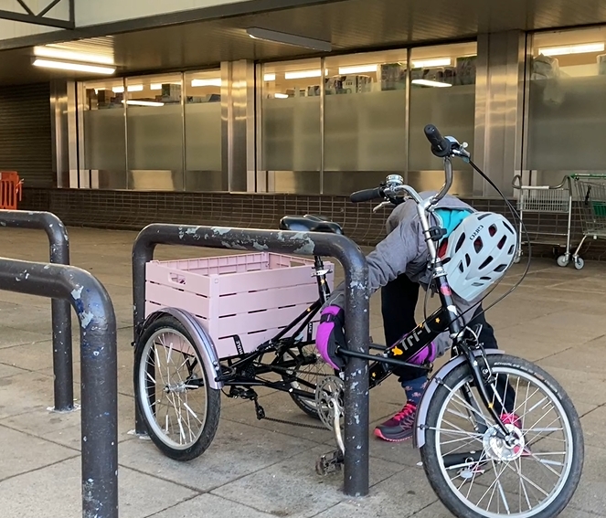 Kid3 is putting a D lock around her trike, which has a big pink crate on the back between the two rear wheels. She’s struggling to position the trike so that she can lock a secure part of the trike onto a battered and unusually fat Sheffield stand.