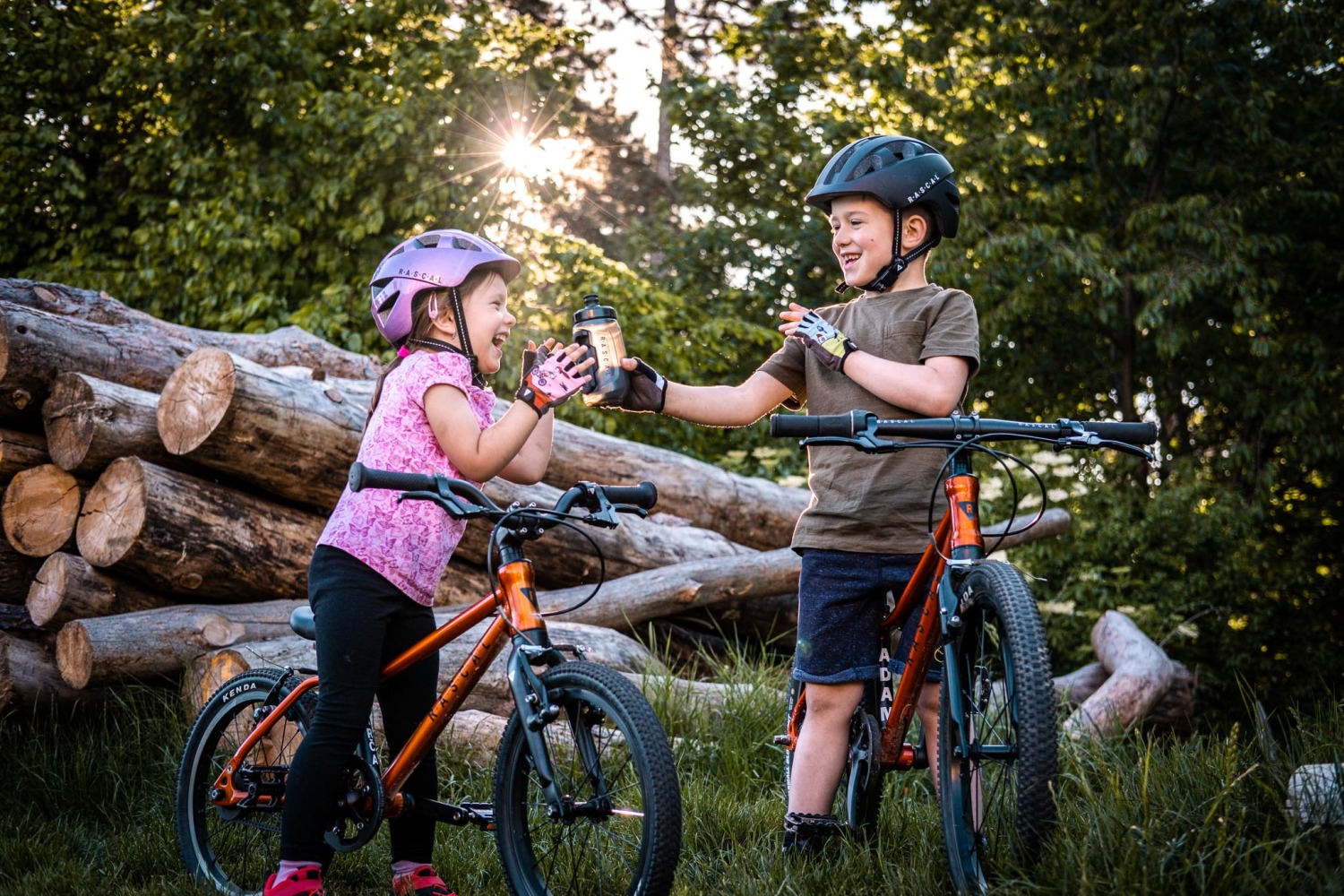 Photo of two children with their Rascal kids bikes in a wood.  The boy is passing the girl a water bottle. They both look happy.  They are both wearing Rascal helmets. The sun is shining through the leaves