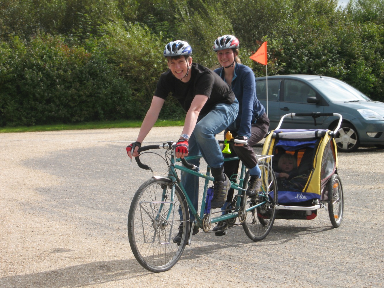 Geoff and Kate riding an old turquoise tourer tandem towing a yellow and blue double child trailer. One child is just visible through the mesh front of the trailer.