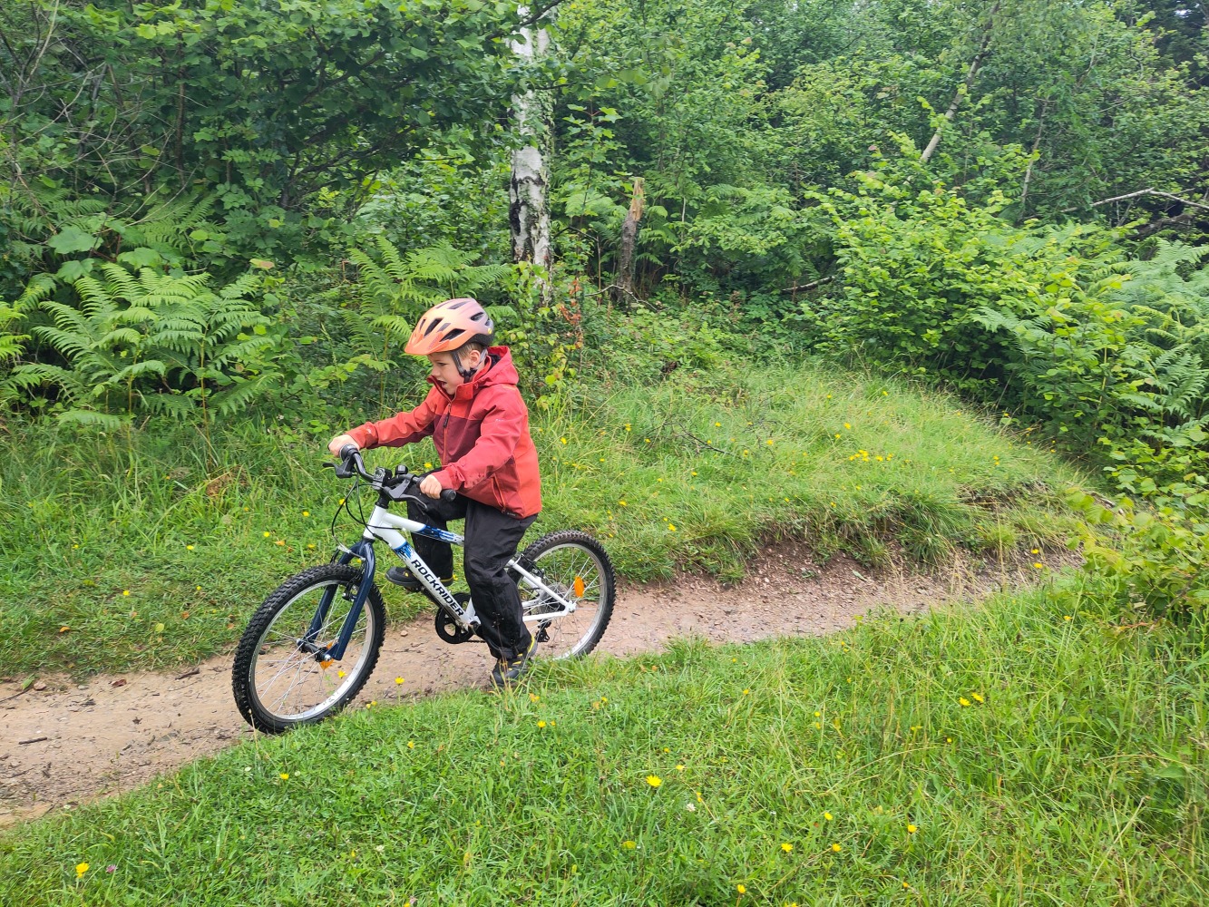 Boy in a red coat riding the Rockrider 20" MTB Expl 120 affordable kids mountain bike along a dirt path 