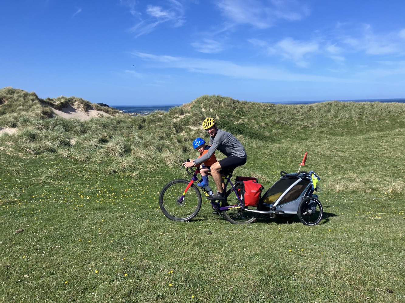A man cycling with his child along a grassy path with a bike trailer on the back of the bike