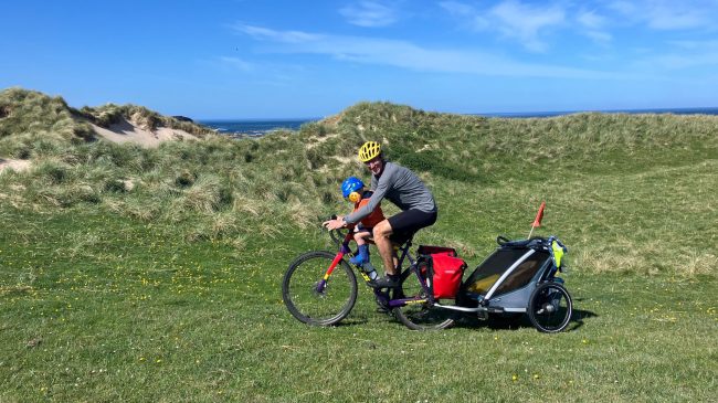 A man cycling with his child along a grassy path with a bike trailer on the back of the bike