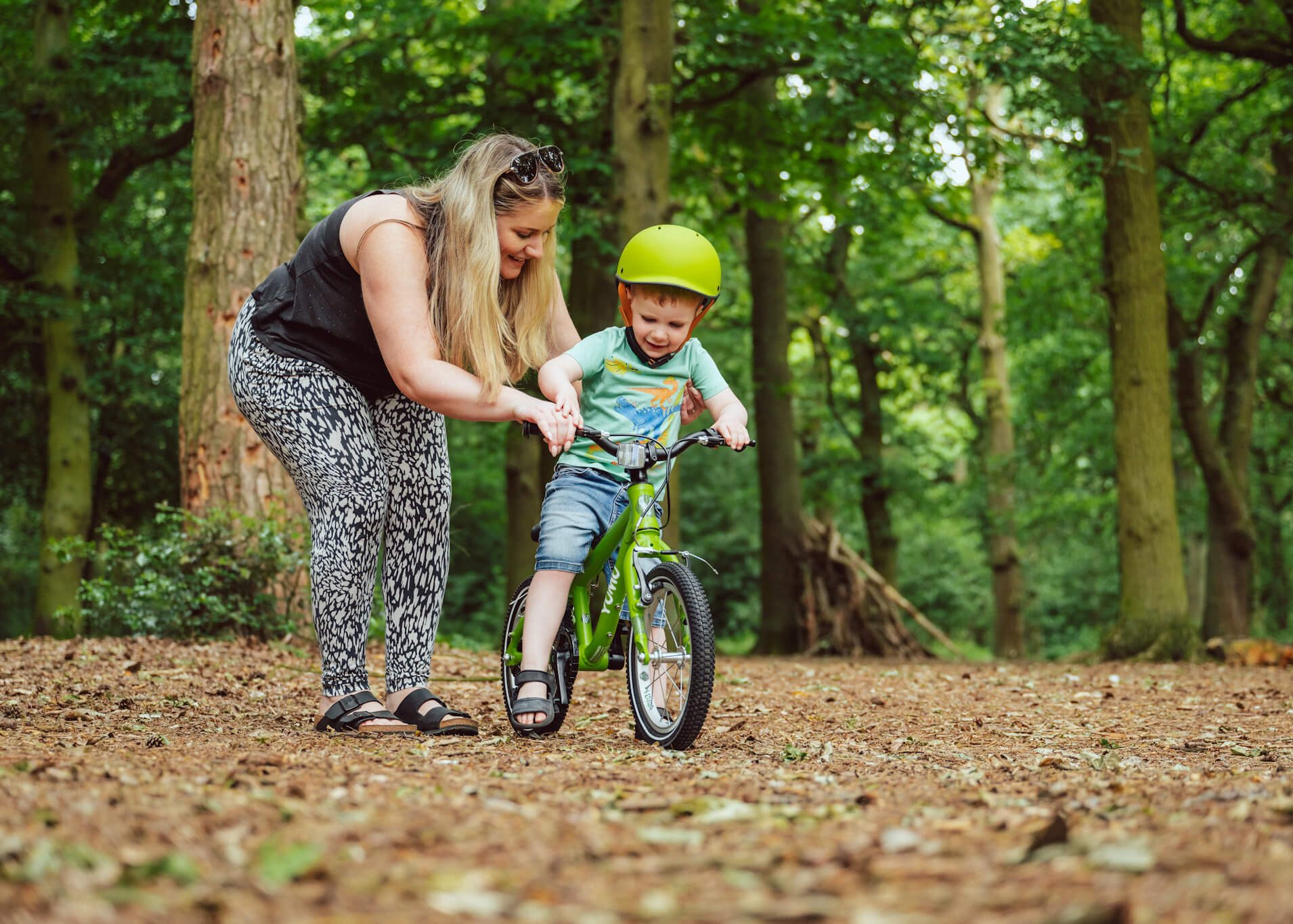 Young boy learning to pedal on a green YOMO bike with his mums help