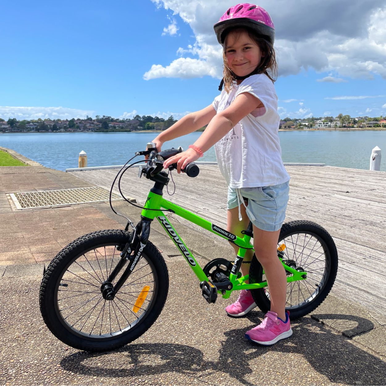 Girl on a large balance bike by Kidvelo at the seaside. Bike is green and girl is about 5 years old. She is wearing a white t-shirt blue shirts and ponk shoes